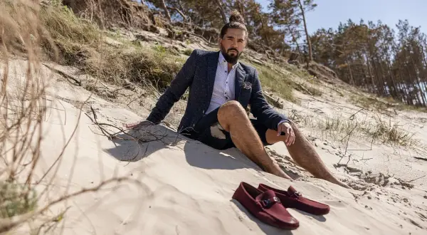 Mokasyny męskie stylizacje na plaży. Mężczyzna w marynarce i szortach siedzi na piasku, obok niego leżą bordowe mokasyny.