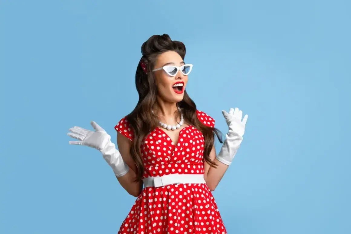 Kobieta w czerwonej sukience w białe grochy, białych rękawiczkach i okularach w stylu pin-up girl stylizacja, z uniesionymi rękami.