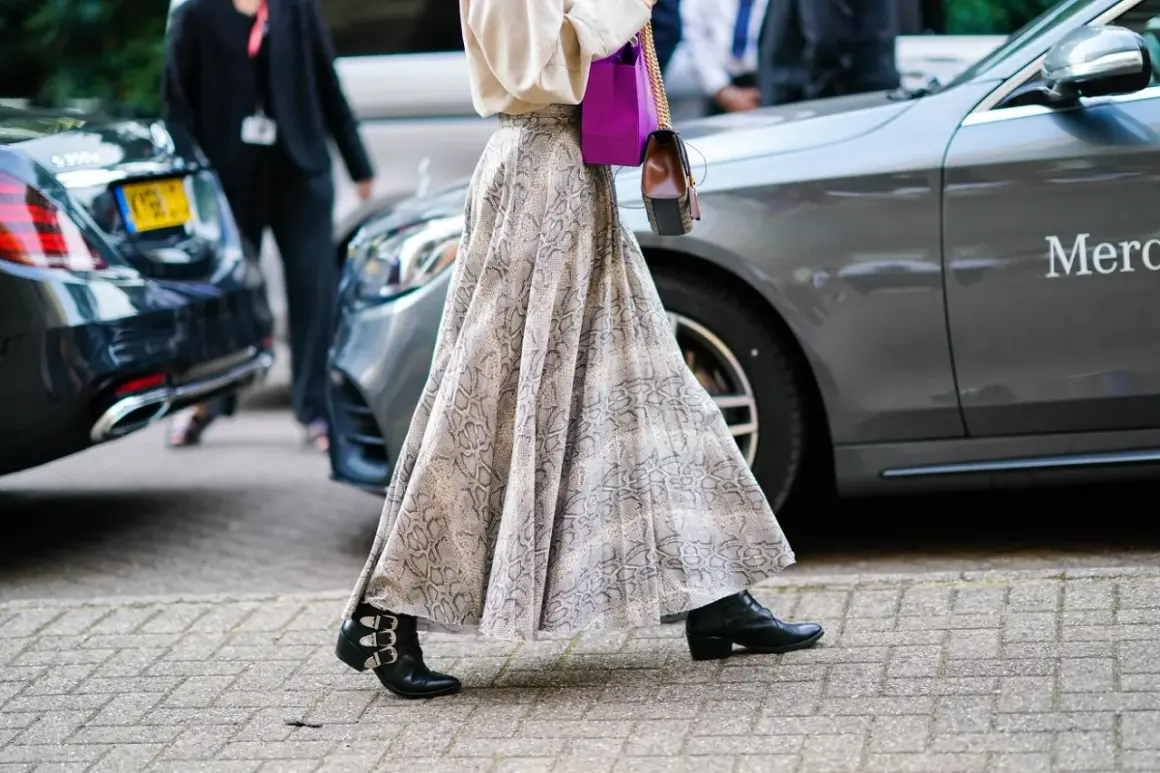 Elegancka długa spódnica w wężowy wzór, zestawiona z botkami i fioletową torebką, tworzy modne stylizacje.