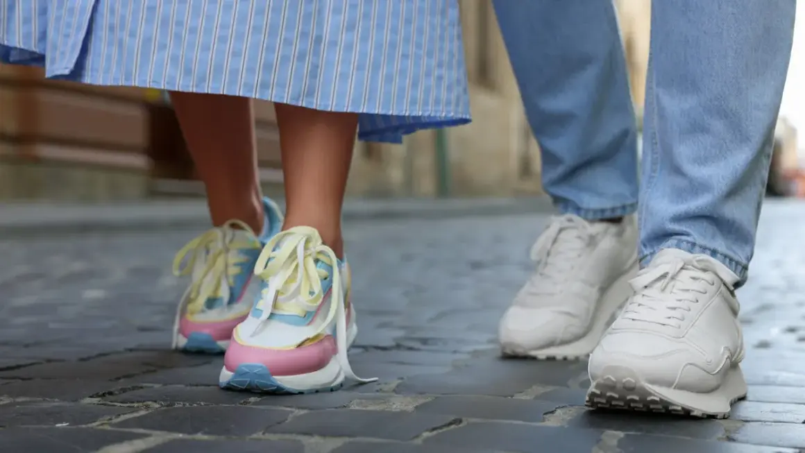 Kolorowe, modne sneakersy 2026 na nogach osoby w sukience i białe na nogach w jeansach, spacerujące po bruku.