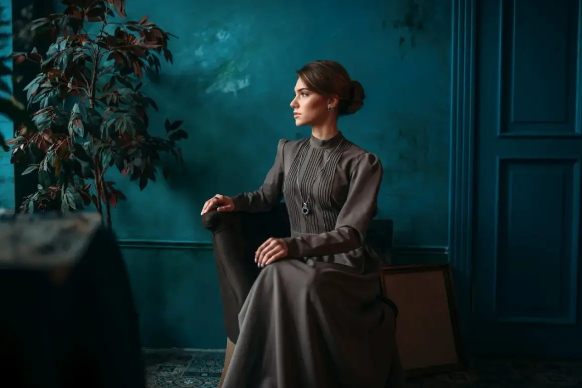 Elegancka kobieta w sukni w stylu light academia siedzi w fotelu, w tle ciemnozielona ściana i roślina.