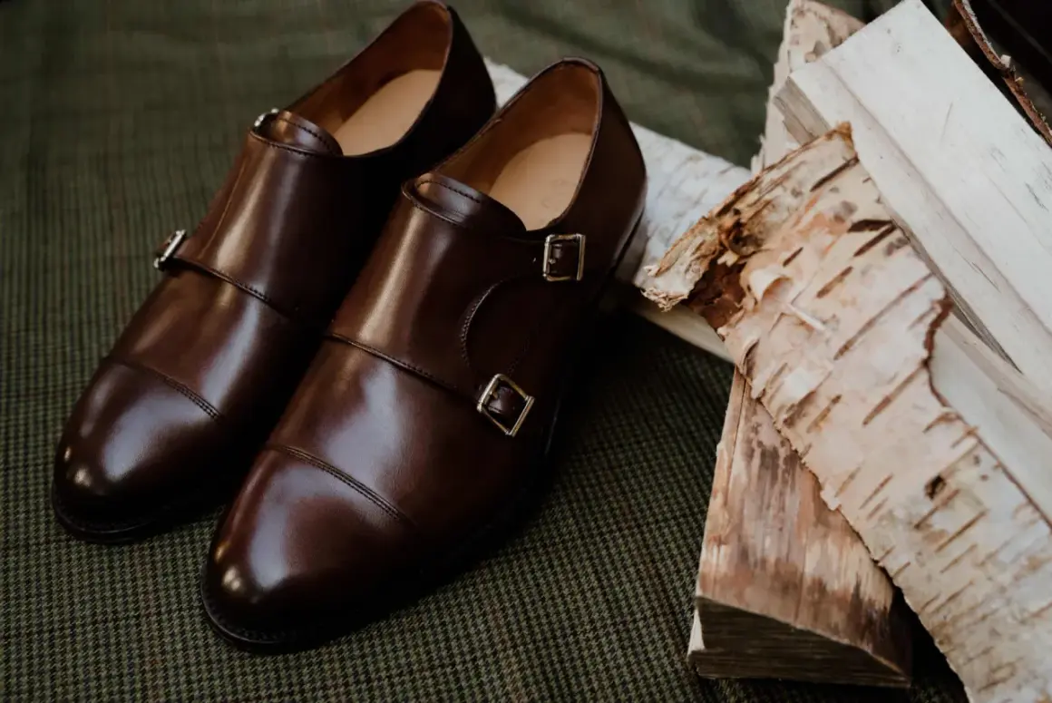 Eleganckie brązowe buty do czarnych spodni, z podwójnymi klamrami, leżą obok polan brzozowych na tweedowym tle.
