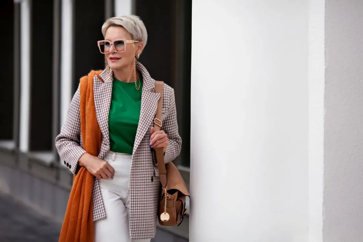 Elegancka kobieta po 60. prezentuje modne stylizacje: marynarka w kratkę, zielona bluzka, białe spodnie, pomarańczowy szal i torebka.