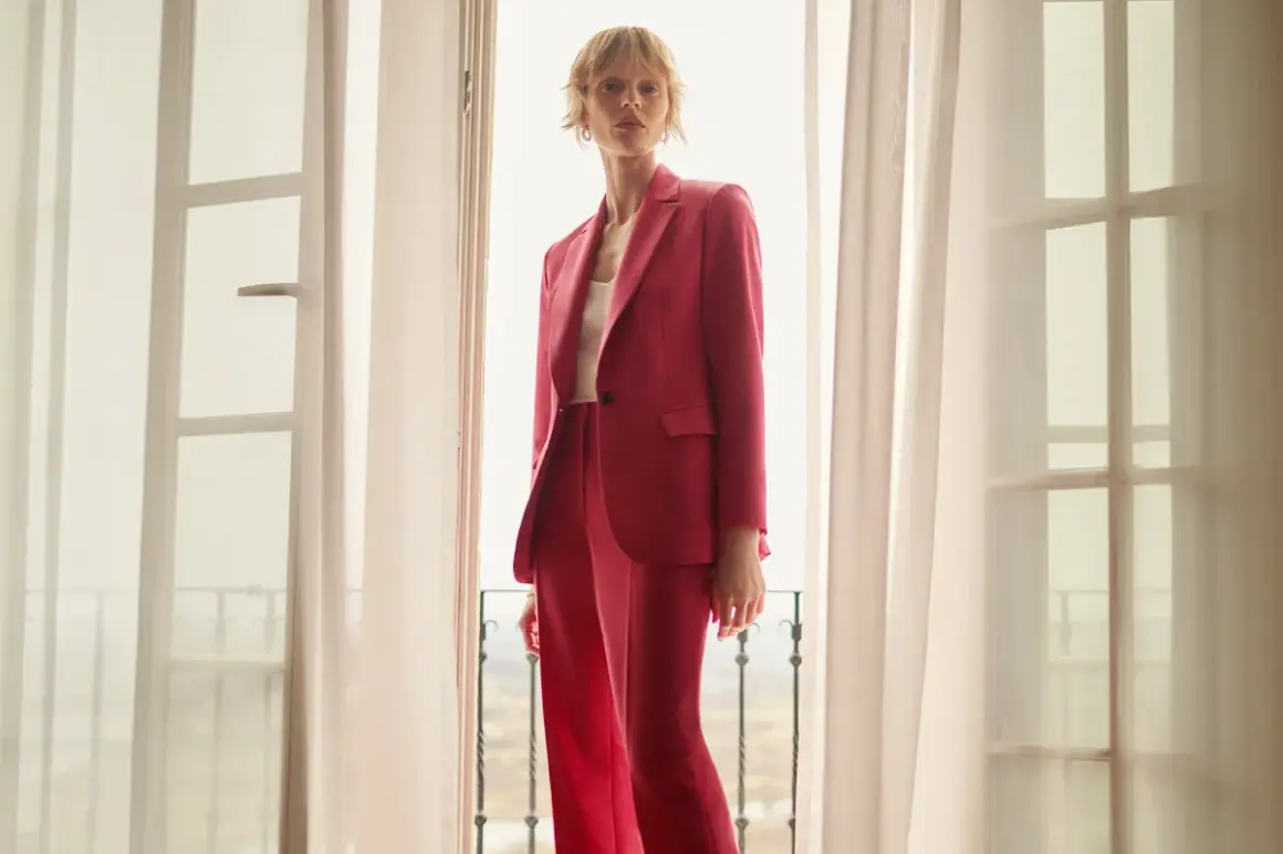 Elegancka stylizacja na chrzciny ze spodniami: kobieta w różowym garniturze stoi przy oknie.