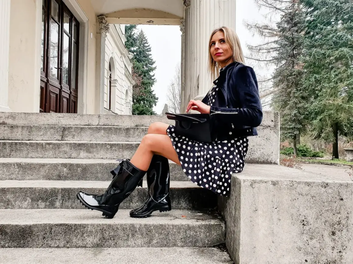 Elegancka kobieta w grochy, w czarnych, błyszczących kaloszach ze skarpetą, siedzi na kamiennych schodach.