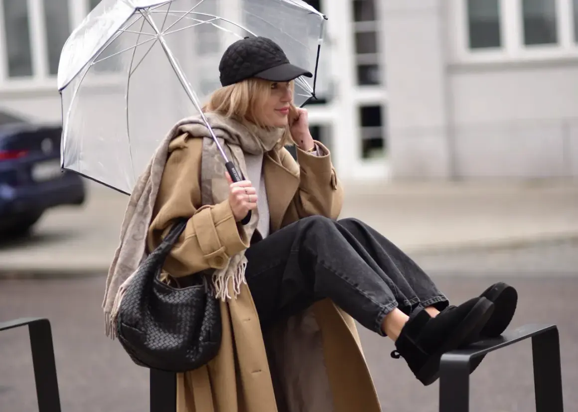 Kobieta w beżowym płaszczu, czapce z daszkiem i jeansach prezentuje modne emu stylizacje, chroniąc się pod przezroczystym parasolem.