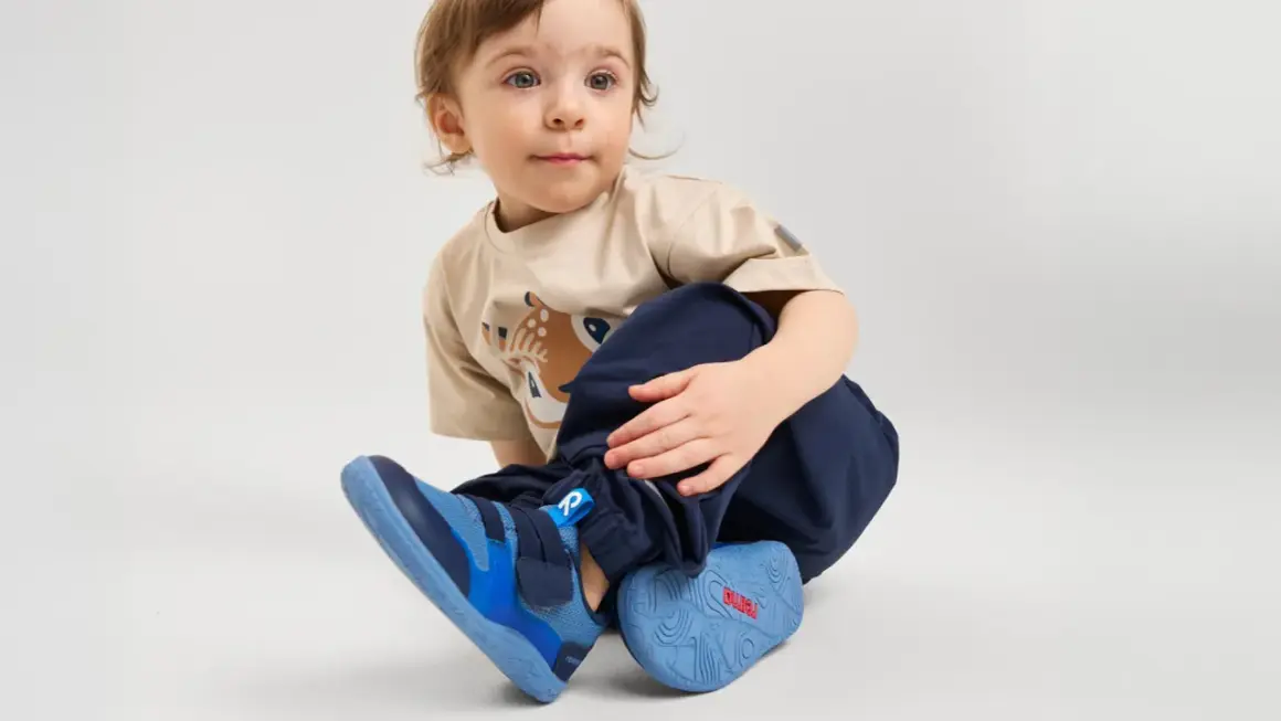 Małe dziecko w beżowej koszulce z jelonkiem i granatowych spodniach, nosi niebieskie buty sportowe.