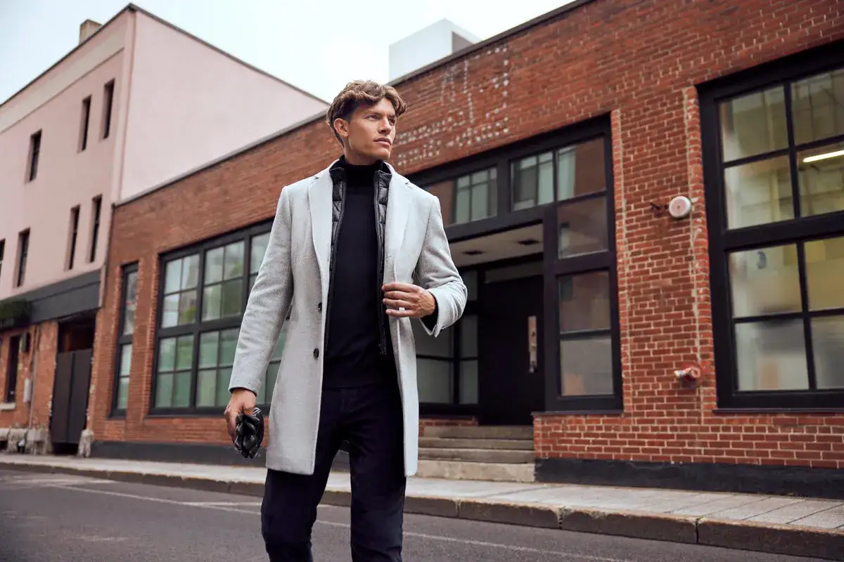 Mężczyzna w eleganckim ubiorze męskim, w jasnym płaszczu i czarnym golfie, idzie ulicą przed ceglanym budynkiem.