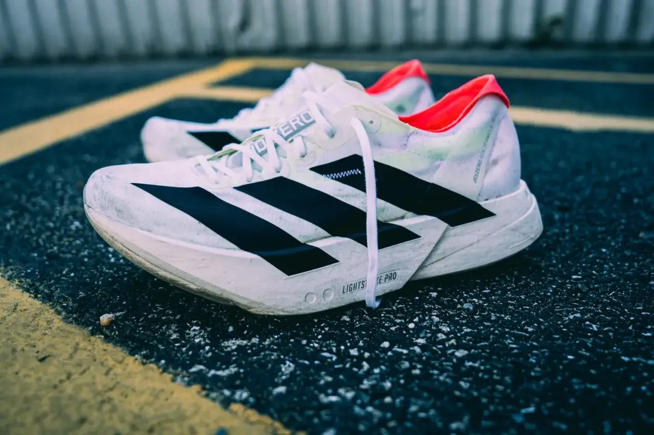 Białe buty do biegania adidas adios pro 4 z czarnymi paskami i neonowym wykończeniem, leżące na asfalcie z ż&oacute;łtymi liniami.