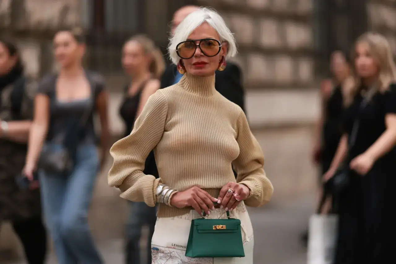 Elegancka pani po 60. w beżowym golfie i spódnicy z mapą. Stylowe okulary i zielona torebka dopełniają jej modną stylizację.