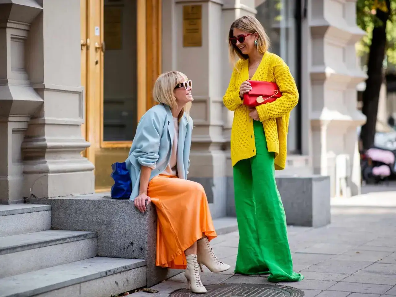 Dwie kobiety prezentują odważny styl color block: jedna w błękitnej marynarce i pomarańczowej sp&oacute;dnicy, druga w ż&oacute;łtym kardiganie i zielonych spodniach.