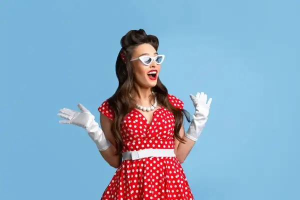Kobieta w czerwonej sukience w białe grochy, białych rękawiczkach i okularach w stylu pin-up girl stylizacja, z uniesionymi rękami.