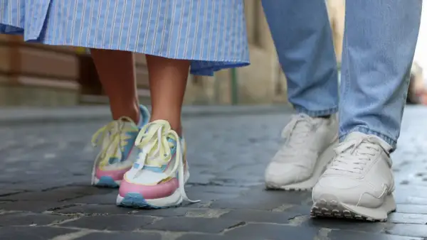 Kolorowe, modne sneakersy 2026 na nogach osoby w sukience i białe na nogach w jeansach, spacerujące po bruku.