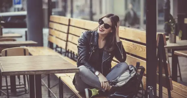 Dziewczyna w skórzanej kurtce i okularach przeciwsłonecznych, z czerwoną szminką, siedzi na ławce w kawiarni. Jej styl to rockowy kobiecy.