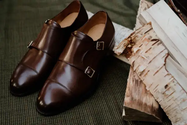 Eleganckie brązowe buty do czarnych spodni, z podwójnymi klamrami, leżą obok polan brzozowych na tweedowym tle.