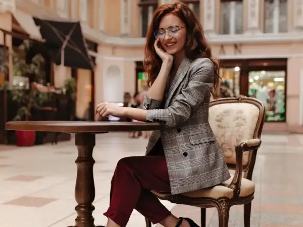 Elegancka kobieta w bordowych spodniach i marynarce w kratę, siedząca przy stoliku. Stylowe bordowe spodnie dodają jej szyku.