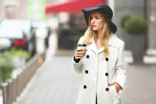 Elegancka kobieta w kapeluszu i białym płaszczu, z kawą w ręku, uosabia paryską modę.