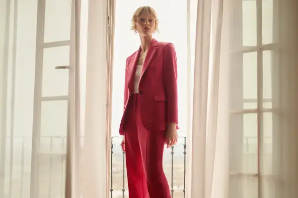 Elegancka stylizacja na chrzciny ze spodniami: kobieta w różowym garniturze stoi przy oknie.