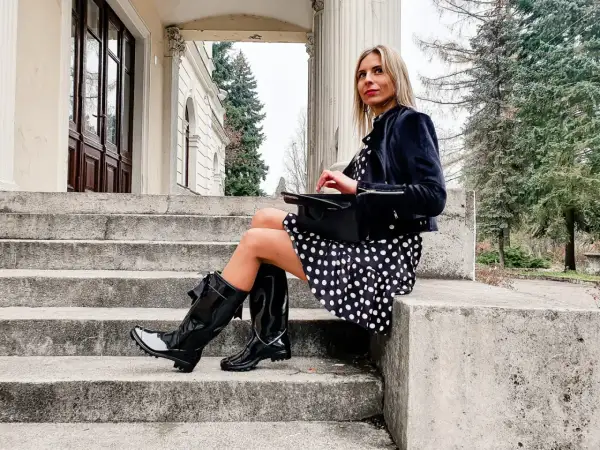 Elegancka kobieta w grochy, w czarnych, błyszczących kaloszach ze skarpetą, siedzi na kamiennych schodach.