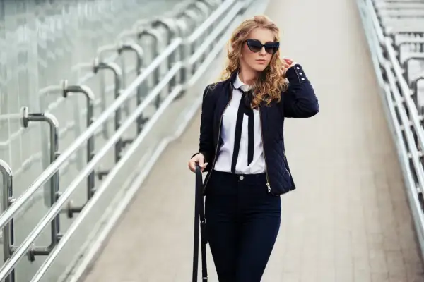 Eleganckie stylizacje z czarnymi spodniami: kobieta w ciemnej kurtce, białej koszuli z kokardą i okularach przeciwsłonecznych idzie po moście.