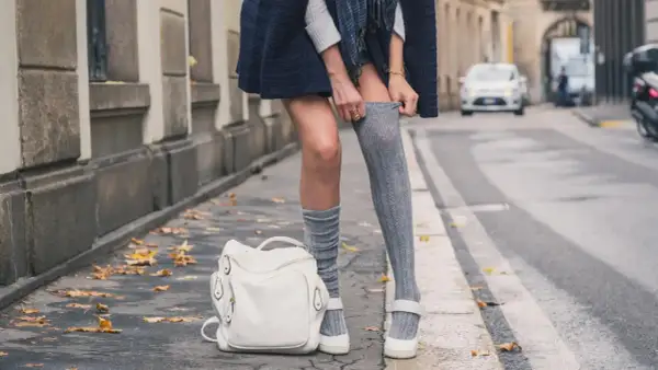 Stylizacja z długimi skarpetkami damskimi: szare, prążkowane, sięgające do połowy uda, zestawione z białą torebką i płaskimi butami.