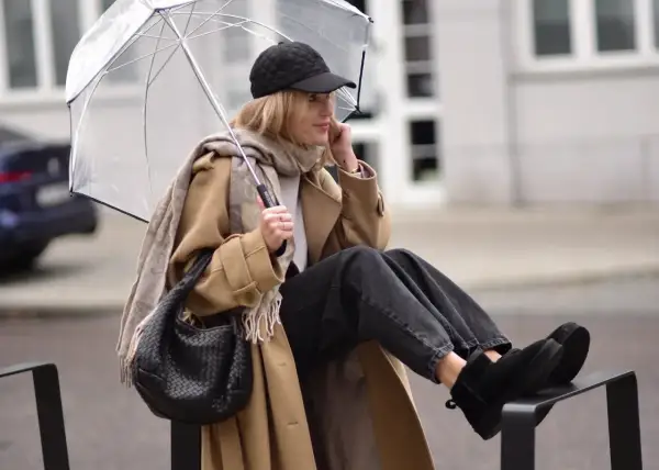 Kobieta w beżowym płaszczu, czapce z daszkiem i jeansach prezentuje modne emu stylizacje, chroniąc się pod przezroczystym parasolem.