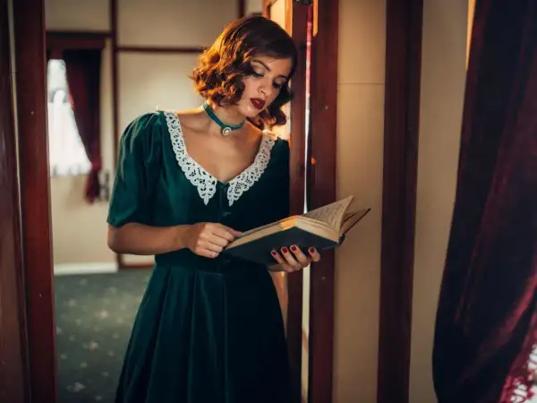 Elegancka kobieta w sukni z lat 40. czyta książkę. Jej stylizacja, z koronkowym kołnierzykiem i biżuterią, idealnie oddaje modę tamtych lat.