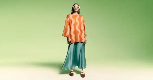 Modelka w okularach w kształcie serc i pomarańczowym swetrze z falistym wzorem. Stylizacja w stylu color block z turkusową, lejącą spódnicą.