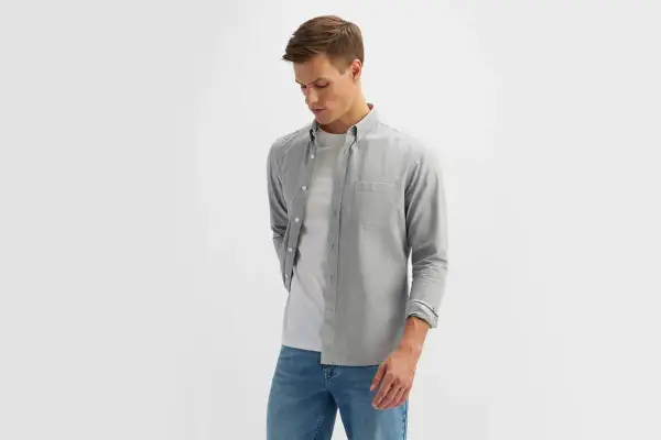 Młody mężczyzna w eleganckim ubiorze męskim: szara koszula z długim rękawem, biały t-shirt i jeansy.